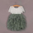 A young girl's party dress with a white lace bodice and short sleeves, paired with a voluminous, multi-layered sage green tulle skirt, hanging on a wooden hanger against a solid mauve background.