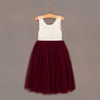 A sleeveless girls' dress hanging on a wooden hanger against a plain taupe-gray background. The dress has a white lace bodice and a full burgundy tulle skirt.
