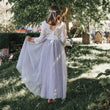A young girl seen from behind wearing a white lace long-sleeve maxi dress with a sheer lavender tulle skirt, holding out the skirt to show its flow in an outdoor grassy garden under a tree.