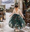A young girl viewed from behind wearing a long-sleeved lace top and a full tulle skirt with embroidered white flowers and green leaves in a snowy rustic courtyard.