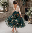 A young girl viewed from behind wearing a floral tulle dress with a lace bodice, standing in a rustic indoor setting with flowers and lanterns.