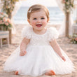 Baby in a white dress sitting outdoors with flowers and a bench in the background