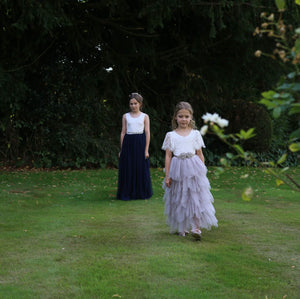Two young girls in formal dresses standing on a grassy area with trees in the background.