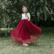 Young girl in a white top and red skirt standing outdoors with greenery in the background