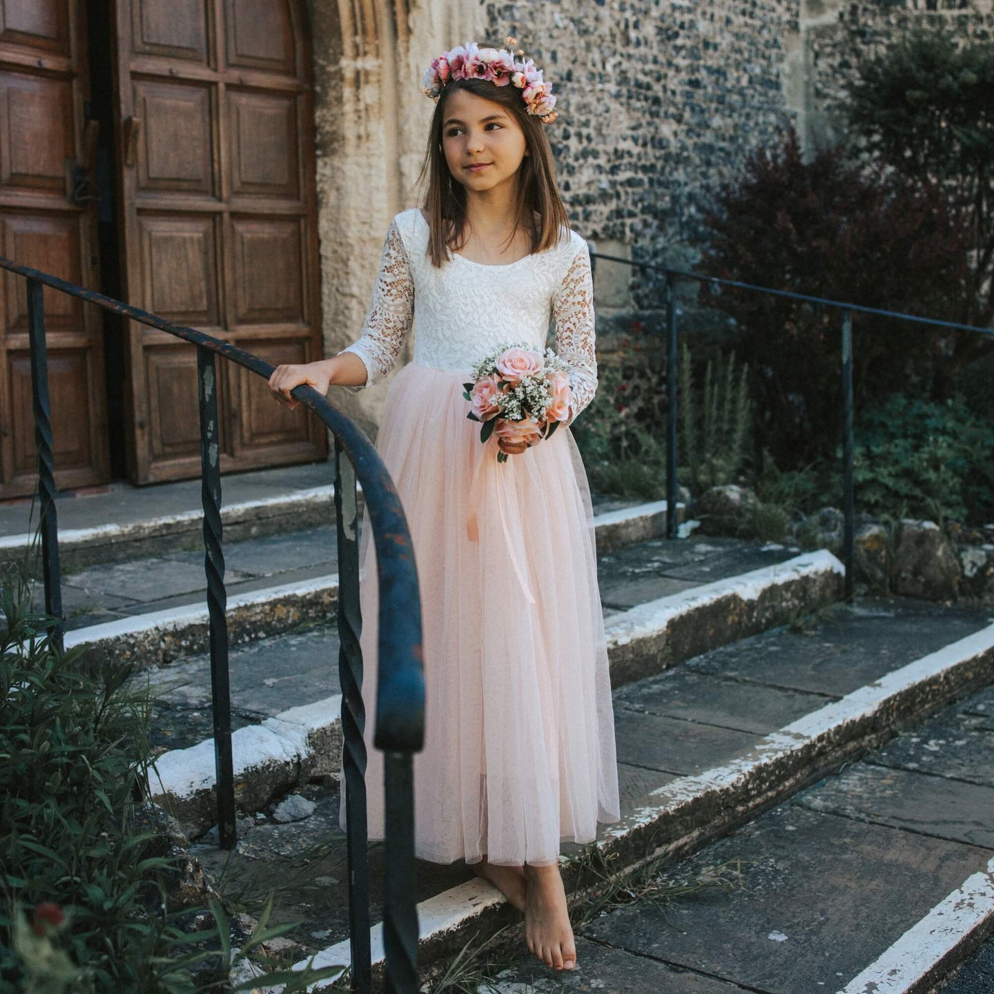 Young girl in a white lace top and BLUSH dress with a flower crown standing on stone steps.