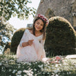 Young girl in a white dress with floral headband sitting on grass with flowers.