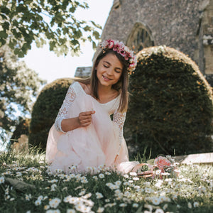 Young girl in a white dress with floral headband sitting on grass with flowers.