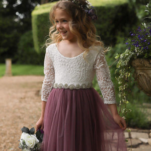 Young girl in a white lace top and purple skirt standing outdoors with greenery in the background
