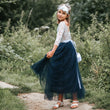 Young girl in a white lace top and blue skirt standing outdoors with greenery around.