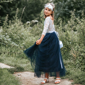 Young girl in a white lace top and blue skirt standing outdoors with greenery around.