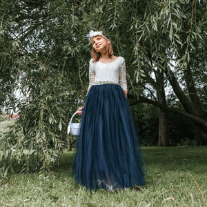 Young girl in a white top and navy blue skirt standing in a grassy area with trees.