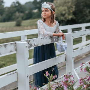 Young girl in a white lace top and blue skirt standing on a white wooden bridge with greenery in the background.