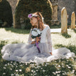 Young girl in a white dress with a floral headband sitting on grass with flowers.