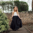 Young girl in a white top and black skirt with a flower crown standing outdoors.