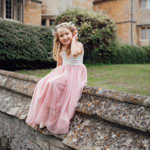 Young girl in a white top and pink dress sitting on stone steps with a building and greenery in the background