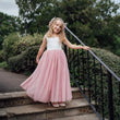 Young girl in a white top and pink skirt dress standing on stone steps with greenery in the background
