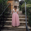 Child in a pink dress with a white lace top standing on stone steps in a garden.