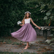Young girl in a purple skirt and floral headband standing outdoors with greenery around.