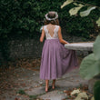 Girl in a purple dress with a floral headband walking away from the camera in a garden setting.