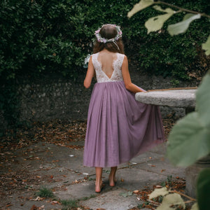 Girl in a purple dress with a floral headband walking away from the camera in a garden setting.