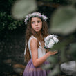 Young girl in a purple dress with a floral wreath and bouquet, standing among greenery.