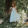 Young girl in a white and light blue dress standing under a tree with greenery around.