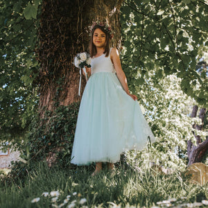 Young girl in a white and light blue dress standing under a tree with greenery around.