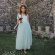 Young girl in a light blue dress holding flowers against a stone wall.