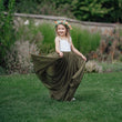 Young girl in a long green dress with a flower crown standing on grass.