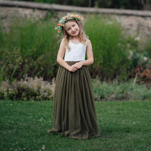 Young girl in a white top and green skirt with a flower crown standing in a grassy field.