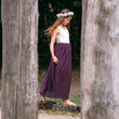 Young girl in a white top and purple skirt standing between two wooden posts in a natural setting.
