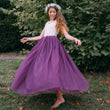 Young girl in a white top and purple skirt standing outdoors with greenery in the background