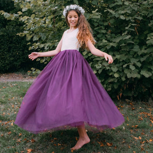 Young girl in a white top and purple skirt standing outdoors with greenery in the background