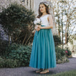 Young girl in a white top and teal skirt standing outdoors with greenery and a building in the background.