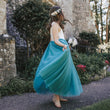 Young girl in a teal blue dress standing in front of a stone building with greenery.