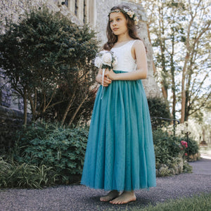 Young girl in a white top and teal skirt standing outdoors with greenery and a building in the background.