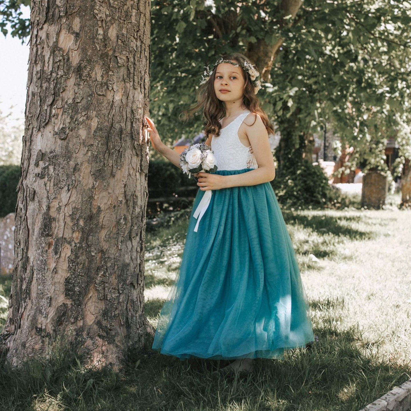Young girl in a white top and teal skirt standing next to a tree in a park.