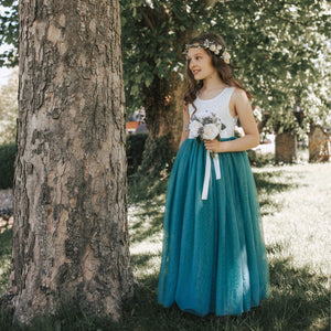 Young girl in a white top and teal skirt standing next to a tree in a park.