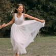 Young girl in a white dress standing outdoors with greenery in the background
