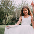 Young girl in a white dress with a tiara, standing outdoors with trees in the background
