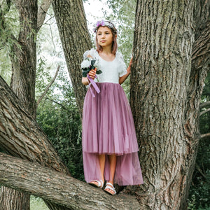 Young girl in a white top and mauve skirt standing between two tree trunks in a forest.