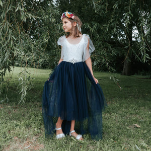 Young girl in a white top and navy blue skirt standing in a grassy area with trees.