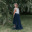 Girl in a white top and navy blue skirt standing in a grassy area with trees.