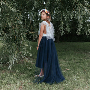 Girl in a white top and navy blue skirt standing in a grassy area with trees.