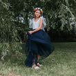 Young girl in a white top and navy blue skirt with a floral crown standing in front of green foliage.