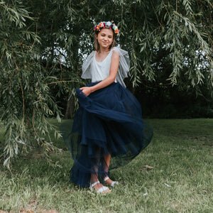 Young girl in a white top and navy blue skirt with a floral crown standing in front of green foliage.