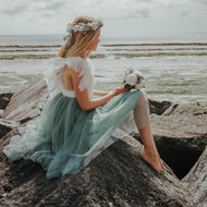Flower girl in a sage green Bohemian Serendipity dress with a floral headband sitting on rocks by the water.
