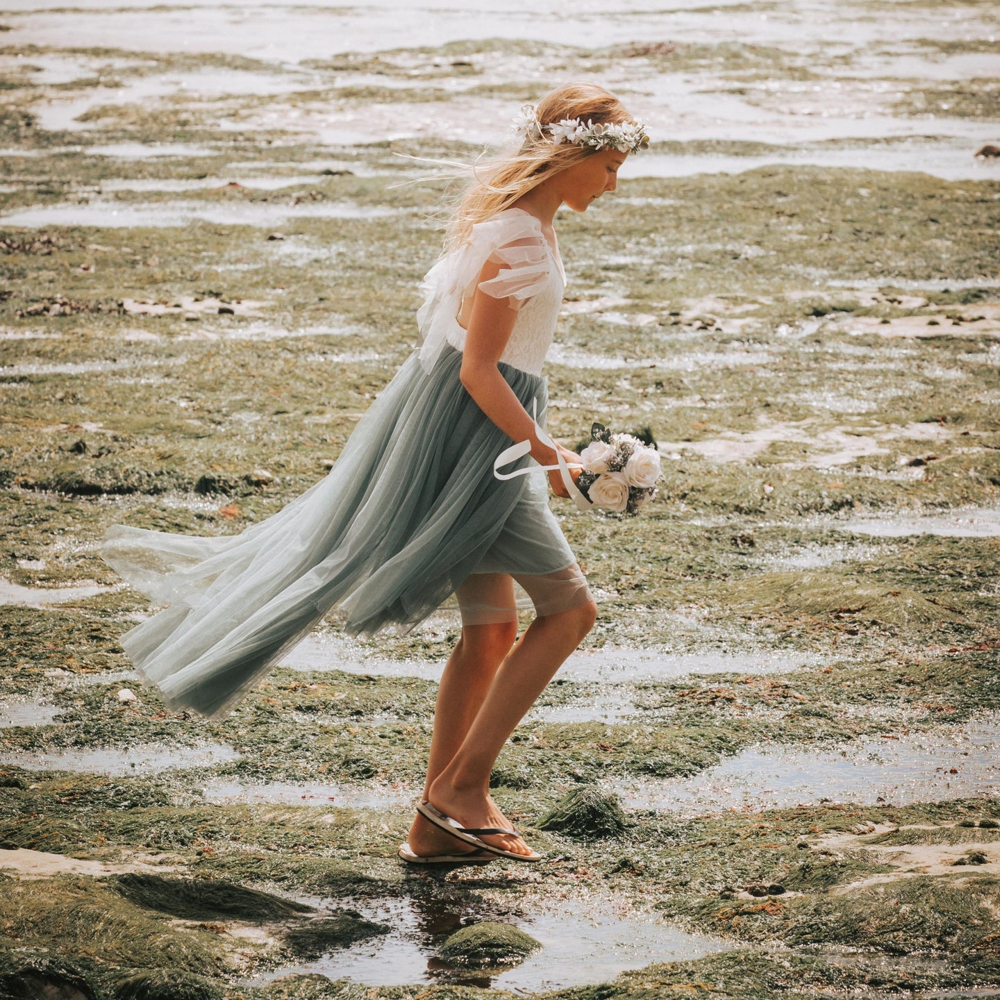 Girl in a flowing dress from UK Flower Girl Boutique walking on a beach with a bouquet.
