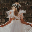 Girl in a white dress with ruffled sleeves standing against a brick wall.