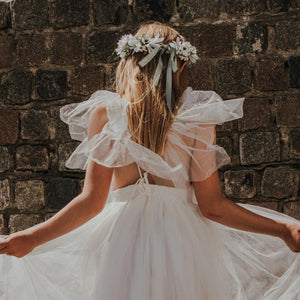 Girl in a white dress with ruffled sleeves standing against a brick wall.